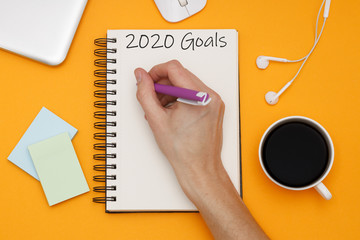 Goals 2020, chek list. Top view of headphones, coffee and notebook with hands on orange background. Flat lay.