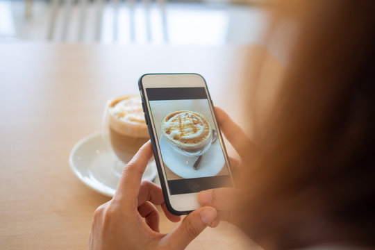 The hand of the woman using a smartphone to take photo of a coffee in a cafe to post on social appication