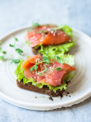Two toasts with salmon slices, sesame, pea sprouts and lettuce on a white ceramic plate on grey background. Copy space