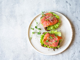 Two toasts with salmon slices, sesame, pea sprouts and lettuce on a white ceramic plate on grey background. Copy space. Top view.