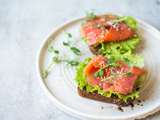 Two toasts with salmon slices, sesame, pea sprouts and lettuce on a white ceramic plate on grey background.