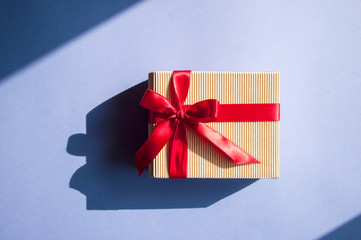 Close up of giftbox tied with bright red color ribbon on purple background in sunlight with shadows.