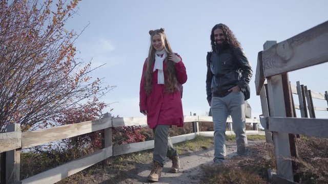 Two biracial people walking down the alley along wooden fence. Positive adult couple dating in the autumn park. Hippie man with long curly hair and attractive blond woman strolling and talking.
