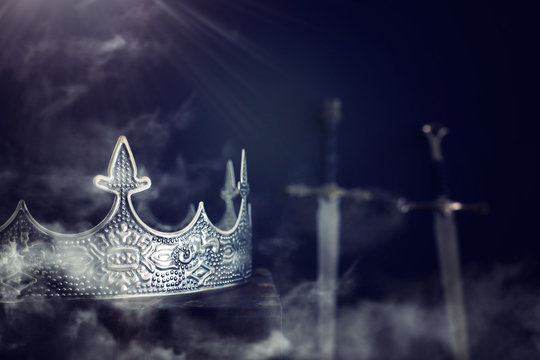 low key image of beautiful queen/king crown over antique book next to sword. fantasy medieval period. Selective focus. mist and fog