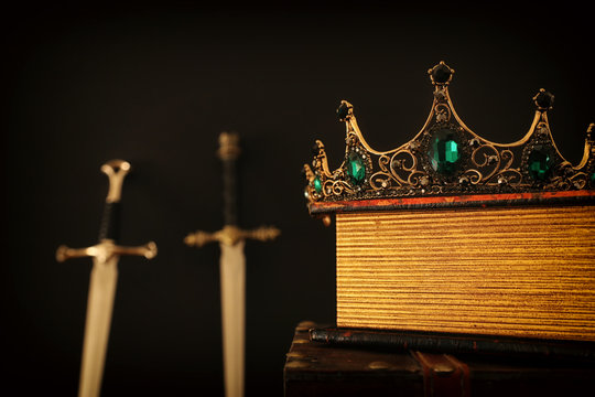 low key image of beautiful queen/king crown over antique book next to sword. fantasy medieval period. Selective focus