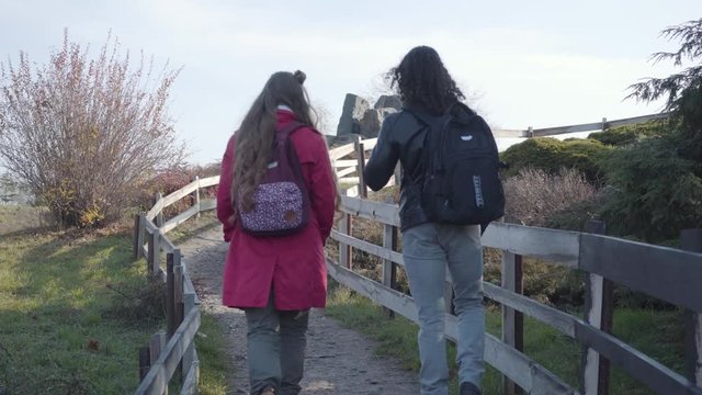 Close-up back view of man and woman in casual clothes walking along wooden fence in autumn park. Positive young multiracial couple dating outdoors. Leisure, free time, weekend activities.