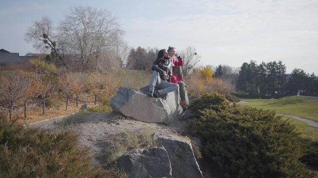 Side view of hippies sitting on rocks in autumn park and talking. Positive carefree couple dating outdoors. Middle Eastern man and Caucasian woman in casual clothes restingafter walk.