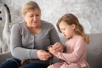Senior grandmother knitting together with her granddaughter at family home. Woman with little girl crochet sitting on a sofa in the living room, life style