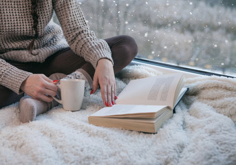 Woman sitting by the window reading book drinking coffee. Winter snowing landscape outside  - 307397803
