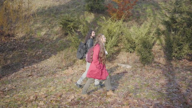 Positive young biracial couple with backpacks walking in the autumn forest. Hippie man and woman in casual clothes strolling outdoors. Dating, love, togetherness, leisure. - Powered by Adobe