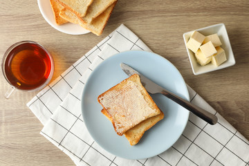 Toasts on plate, towel, tea and butter on wooden background, top view