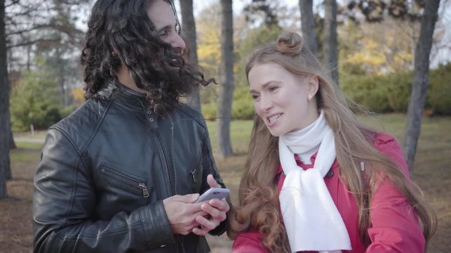 Funny biracial couple listening rock in autumn park. Man with long curly hair and beard and pretty woman listening to songs in headphones. Cheerful boyfriend and girlfriend having fun together
