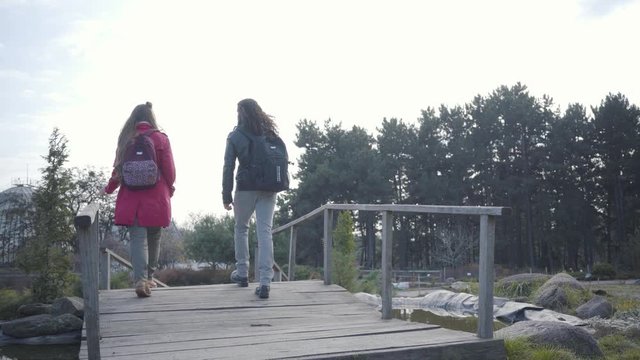 Back view of Caucasian woman and Middle Eastern man strolling along wooden alley in autumn park. Hippie couple in casual clothes walking together and talking. Romantic date outdoors.