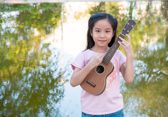 Little asian child girl play the ukulele, in the garden near the pond