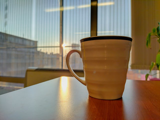 Closeup of white mug cup made of porcelain ceramics placed on a wooden desk with beautiful view of outside through the mirror while at a job