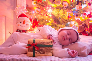 Baby asleep on a cushion dressed in white, lights and warm Christmas decorations, hugs golden gift