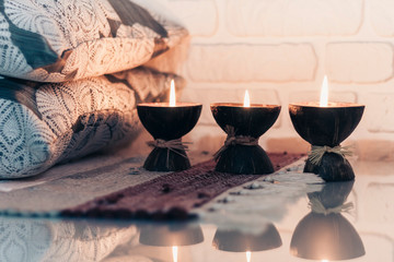 Burning spa aroma candle in coconut shell on a knitted multicolored rug and gray pillows opposite the white wall, cozy home interior.