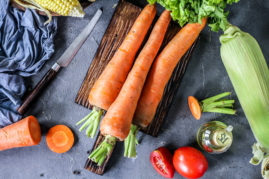 Photo of Fresh Carrots On Wooden Cutting Board. On wooden Dark Background. Slice of carrots with green leaves. Carrot around vegetables, salt, black pepper, corn, broccoli. Top View. Image