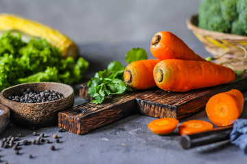 Photo of Fresh Carrots On Wooden Cutting Board. On wooden Dark Background. Slice of carrots with green leaves. Carrot around vegetables, salt, black pepper, corn, broccoli. Drops of water. Image