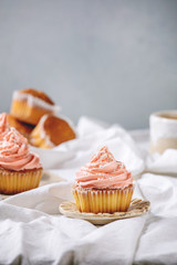 Homemade cupcakes with pink buttercream and coconut flakes served with ceramic cup of tea on white folded tablecloth.