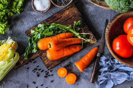 Photo of Fresh Carrots On Wooden Cutting Board. On wooden Dark Background. Slice of carrots with green leaves. Carrot around vegetables, salt, black pepper, corn, broccoli. Drops of water. Image