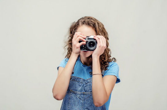 Curly brunette girl in denim overalls is holding a vintage film camera in her hands. Takes a picture