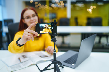 Hand woman holding camera for recording video while sitting at office. Teenager student in a yellow...