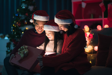 Young Asian family celebration in Christmas day, father mother and daughter siting on sofa and open gift box which exciting smiling and felling happy in living room at home at night time. Merry Xmas.