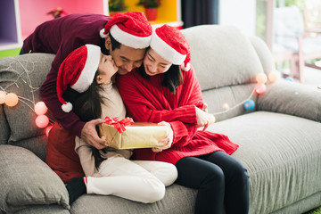 Young Asian family celebration in Christmas day, parent and daughter siting on sofa which hold gift box, girl kiss father which smiling felling happy in living room at home morning time. Merry Xmas.