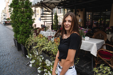 Happy brunette young girl in black t-shirt sitting on a street terrace of a cafe decorated with flowers and looking at the camera