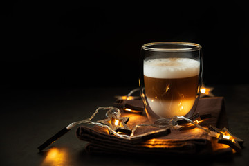 Front view on thermo glass cup of latte coffee decorated Christmas lights on dark wooden table. Morning beverage.  Christmas and New Year holidays concept