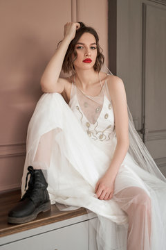 Provocative woman with red lips in white wedding dress and black brutal shoes sitting on chest of drawers leaning on hand and looking at camera against wall of modern apartment