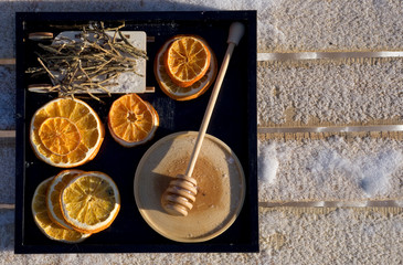 Winter flat lay with dry orange, honey with wooden spoon, toy sled with Rhododendron adamsii  on black tray    on snow