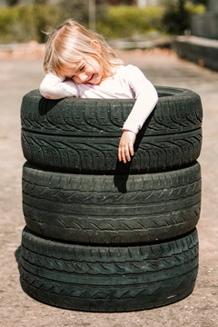 Happy adorable little girl resting in stack of car tires while having fun and playing outdoors on summer day