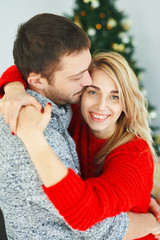 Romantic couple in love embracing, kissing near the Xmas tree at home. Happy young family celebrating Christmas and New Year, happy time together. Pretty woman looking at camera