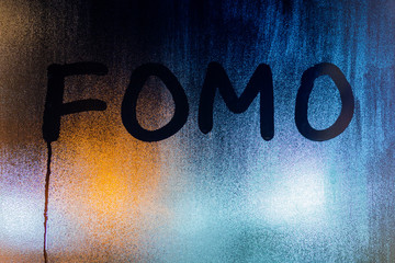 the word fomo- fear of missing out - written on night wet window glass with blurred background