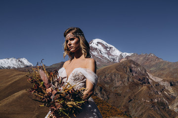 Fototapeta na wymiar young and attractive woman dressed in wedding dress in boho style posing for photo on mountains background, perfect makeup, hairstyle and wedding floristry, happy bride in anticipation of groom