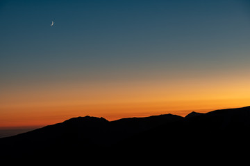 Sunset over silhouetted mountains with a crescent moon rising 