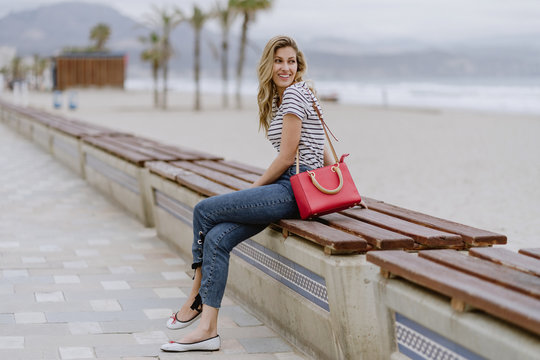 Attractive modern long haired blonde female with beautiful smile wearing casual striped shirt looking away while sitting at bench - Powered by Adobe