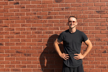 Happy attractive young man in sunglasses, dressed in black t-shirt standing with hands on a belt, red brick wall on the background