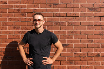 Happy attractive young man in sunglasses, dressed in black t-shirt standing with hands on a belt, red brick wall on the background