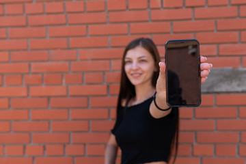 Attractive girl in black top standing near the red brick wall holding phone in hand and shows the screen of the phone in the camera