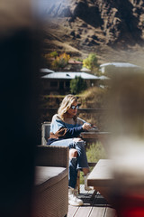 photo through window: stylish attractive girl dressed in warm clothes sits on the terrace of a mountain cafe and lonely smokes a cigarette in warm sunny weather