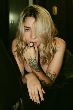 Seductive inviting fair haired tattooed woman in black bra and jeans sitting on sofa with legs spread wide