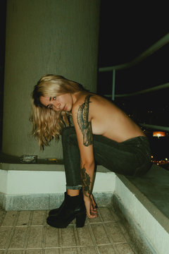 Sexy blonde tattooed female in top less and jeans sitting with legs spread on balcony