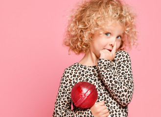 Cute blonde kid girl with curly hair in leopard print sweater is posing with big lollipop, holding her finger at nose