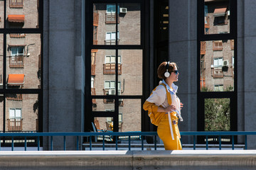 Side view of trendy happy female in wireless headphones and bight yellow clothes with backpack walking against facade windows with reflection of modern building while listening to music in sunlight