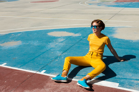 High angle of frowning cool female teenager in yellow clothes with sunglasses while sitting alone on blue basketball sports ground against blurred modern multistory buildings in summer