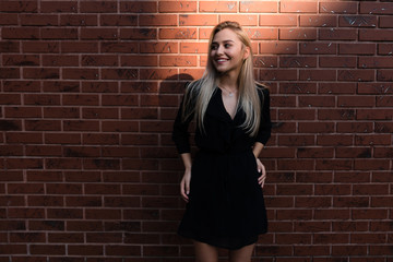 Fashion blonde woman in black dress smiling and looking away, red brick wall on the backgroud