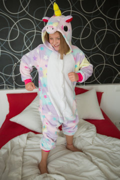 Happy joyful girl in funny kigurumi unicorn pajama playing in bedroom and jumping on bed with red and white bedding